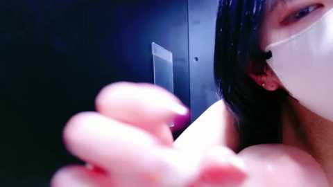 Media: A close-up video of a woman with long black hair, wearing a white mask, holding her breasts, with a dim, blue-lit background.