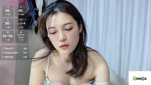 Media: Video of a young Asian woman with fair skin, dark hair, and closed eyes, wearing a white spaghetti-strap top, sitting in a room with hanging clothes and a window.