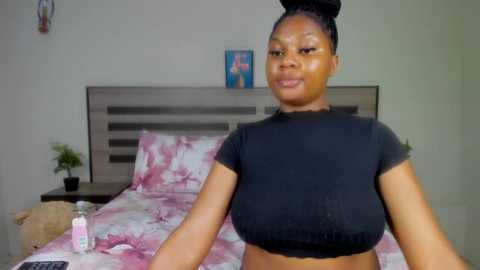 Media: Video of a Black woman with medium skin tone, braided hair, and black crop top, sitting on a bed with pink and white patterned bedding. The room has a wooden headboard, a small potted plant, and a framed picture on the wall.