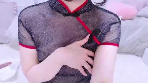 Media: Video of a woman in a sheer, black cheongsam with red trim, wearing it over a white blouse. She's indoors, seated on a light-colored couch, with a soft, pastel background.