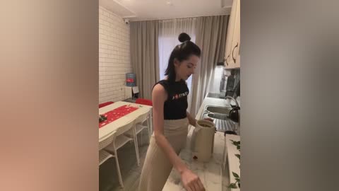 Media: Video of a woman with a high ponytail, wearing a black crop top and beige pants, washing dishes in a small, modern kitchen with white subway tiles and gray curtains.