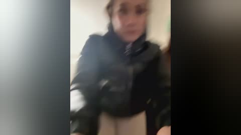 Media: A blurred video shows a woman with fair skin, wearing a black puffer jacket, standing indoors with a neutral background. Her face is slightly out of focus.