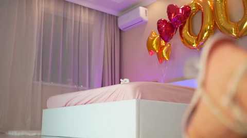 Media: Video of a modern bedroom with a white bed, pink sheets, heart-shaped gold and red balloons, sheer curtains, and an air conditioner.