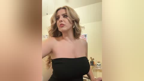 Media: Video of a curvaceous woman with wavy blonde hair, wearing a black strapless dress, standing in a dimly lit room with cream-colored walls.