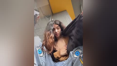 Media: Video of a young woman with long, wavy brown hair, wearing a brown top and blue jeans with eye patches, sitting on a tiled floor with a yellow wall in the background.