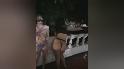 Media: Video of two women in provocative lingerie, standing close together, one wearing a sheer top, the other a black crop top, outdoors at night.