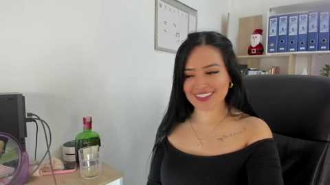 Media: Video of an attractive Asian woman with long black hair, wearing a black off-shoulder top, smiling in an office setting with a whiteboard, bottles, and a Christmas ornament.