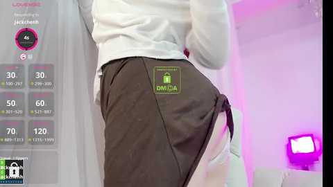 Media: Video of a person in a white long-sleeved top and black shorts with an \"AMAZON\" logo, standing indoors with a digital thermometer display and a purple light in the background.