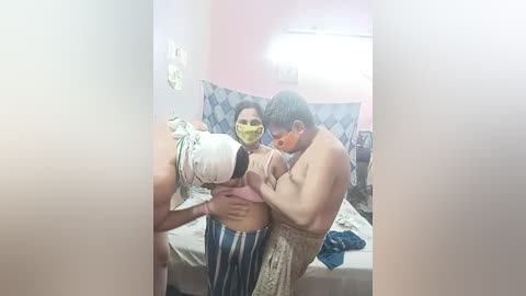 Media: A video shows a shirtless Asian man with glasses and a yellow mask, lifting a shirtless Asian woman with a mask and blue-striped pants in a cluttered bedroom.