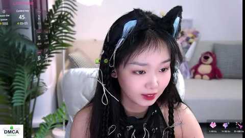 Media: Video of a young Asian woman with long black hair, wearing black headphones and cat ears, seated indoors with a plush toy in the background.