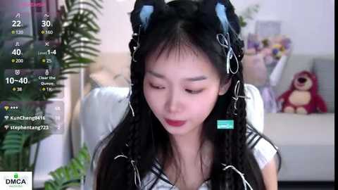 Media: Video of an Asian woman with long black hair, wearing black cat ears, red lipstick, and a white top, sitting in a modern living room with a stuffed toy bear.