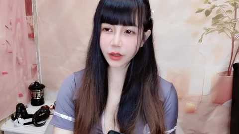 Media: Video of an Asian woman with long, straight black hair and bangs, wearing a light blue top. She has fair skin and is smiling slightly. Background features a soft, beige wall with a potted plant and a black bottle.