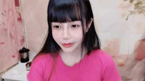 Media: Video of a young Asian woman with long black hair and fair skin, wearing a bright pink top. Background features pastel-colored walls and a potted plant.