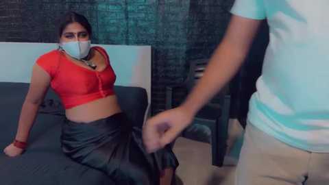 Media: A video depicts a woman in a red crop top and black skirt tied up by a man in a teal shirt, in a dimly lit room with a dark wall and wooden floor.