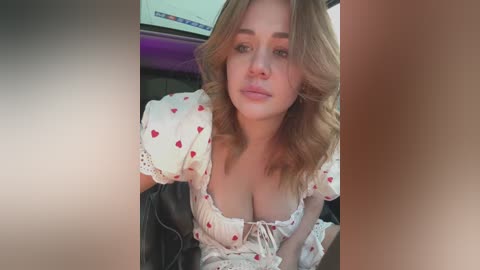 Media: A video of a young Caucasian woman with fair skin and shoulder-length blonde hair, wearing a low-cut, white blouse with red heart patterns, sitting in a car.