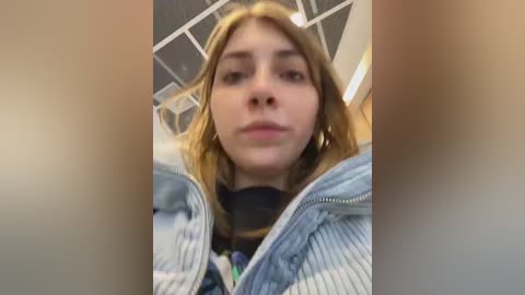 Media: Video of a young woman with light skin, long blonde hair, and wearing a striped jacket. The background features a modern office ceiling with square tiles.