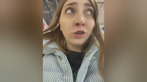 Media: Video of a young woman with light skin, brown hair, and a shocked expression, wearing a striped jacket. The background is blurry, suggesting an indoor setting.