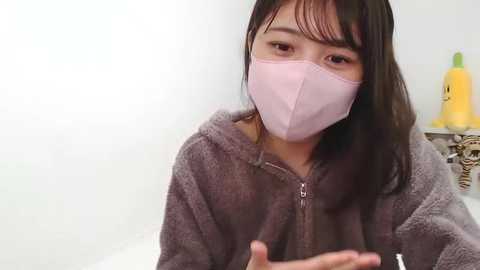 A video of an East Asian woman with straight, shoulder-length dark hair, wearing a light pink face mask and a grey fuzzy hoodie, seated indoors.