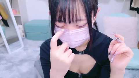 Media: Video of an East Asian woman with straight black hair, wearing a pink face mask and a black top, applying nail polish. Background includes a light teal ottoman, white walls, and a mirror.