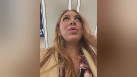 Media: A video of a blonde woman with long hair, wearing a beige jacket and plaid shirt, looking upwards with a thoughtful expression. Background shows a subway car with white ceiling panels.