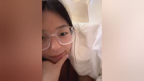 Media: Video of an Asian woman with long black hair, wearing glasses and a white shirt, lying on a beige pillow, partially obscured by a blurred object.