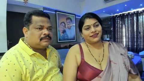 Media: Video of a middle-aged South Asian couple in a modern living room. The man wears a yellow shirt, the woman a pink sari, both smiling. Background includes a framed photo, dark curtains, and blue lighting.