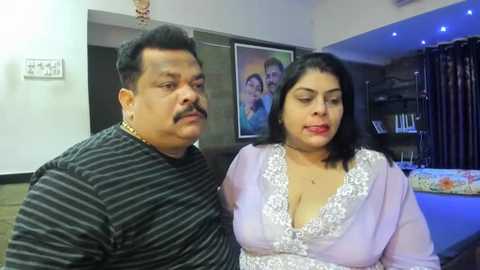 Media: Video of an Indian couple in a modern living room. The man, with a mustache, wears a striped shirt, and the woman, with long black hair, a light pink top with lace detailing. Background includes a framed photo, bookshelf, and a pool table.