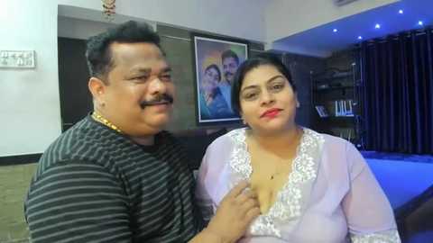 Media: Video of a middle-aged Indian couple in a modern living room; man in striped shirt, woman in white lace top. Background features a framed family portrait, dark curtains, and a pool table.