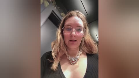 Video of a fair-skinned woman with blonde hair, wearing glasses and a black top, with a chunky silver necklace. She has a surprised expression.