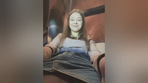 Video of a young woman with long, straight, light brown hair, wearing a light gray shirt, a brown sweater, and a high-waisted denim skirt, seated on a wooden bench in a rustic, warmly lit room with orange walls.