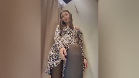 Video of a young woman with fair skin and straight brown hair, wearing a floral-patterned dress and black tights, leaning against beige walls in a narrow hallway.