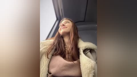 Media: Video of a smiling young woman with long brown hair, wearing a light beige fur-trimmed coat and pink ribbed sweater, looking upwards in a bright, modern interior.