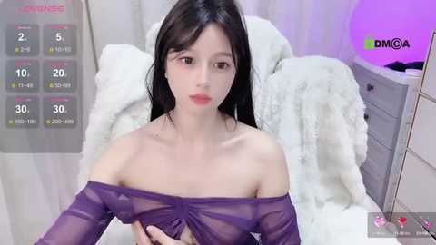 Media: Video of an East Asian woman with long black hair, wearing a sheer purple top, sitting on a white bed. Background includes a white blanket and a purple light.