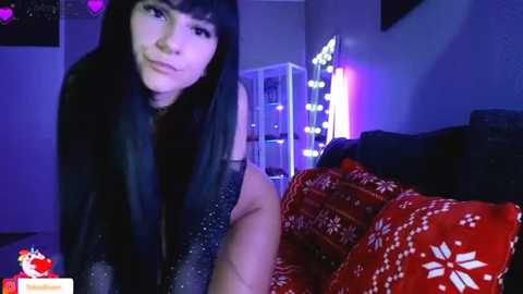 Media: Video of a young woman with long black hair, wearing a black sparkly top, sitting on a bed with red and white patterned blankets, in a dimly lit room with purple lights and a mirror.