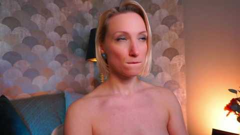 Video of a pale, blonde woman with short hair, wearing blue earrings and no shirt, against a backdrop of textured, abstract wallpaper and a lit lamp.