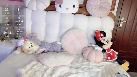 Media: Video of a cozy bedroom with plush toys including a Minnie Mouse doll, a plush cat, and large white pillows. Background features a wooden door and white bedding.