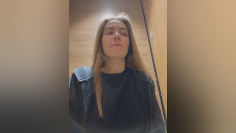 Video of a young Caucasian woman with long, straight light brown hair, wearing a black turtleneck and a denim jacket, looking contemplative in a narrow hallway with beige walls and wooden floor.