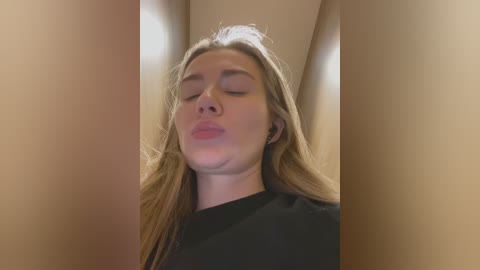 Video of a young Caucasian woman with long, straight blonde hair, closed eyes, and puckered lips, wearing a black shirt, against a beige, indoor background with soft lighting.