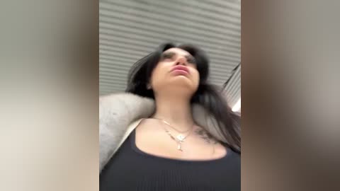 Media: Video of a young woman with long dark hair, wearing a black tank top, silver necklace, and a grey cardigan. She has light skin and is looking upwards, possibly in an airport terminal.