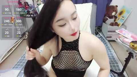 Media: A video of an East Asian woman with long black hair, wearing a black fishnet top, red lipstick, and a white bedsheet, in a cluttered bedroom with a teddy bear and a framed picture.