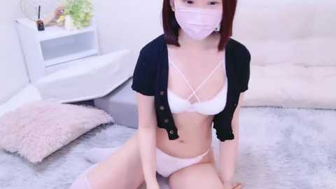 Media: Video of an Asian woman with straight, shoulder-length brown hair, wearing a pink face mask, white lingerie, and a black cardigan, kneeling on a gray carpet in a modern, minimalist bedroom with white walls and a plush white bed.