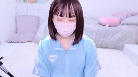 Media: Video of a young Asian woman with straight brown hair, wearing a light blue jacket with a heart emblem, pink mask, and seated on a white floor in a pastel-colored, minimalist bedroom with a bed, pillows, and a dresser.