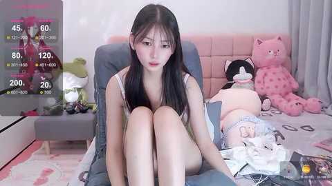 Media: Video of a young East Asian woman with long black hair, sitting on a bed, wearing a white tank top, surrounded by plush toys, in a cozy, girlish bedroom.