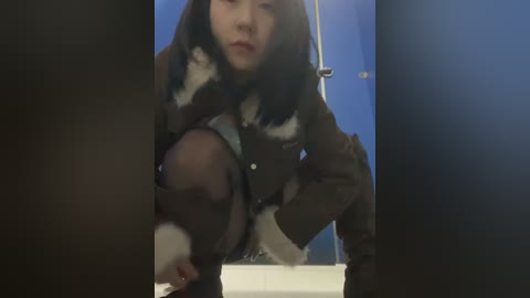 Media: Video of a person in a brown, fur-trimmed jacket, squatting in a blue bathroom, holding a dog's head. The image is dark, with soft lighting, and a blurred, shadowy background.