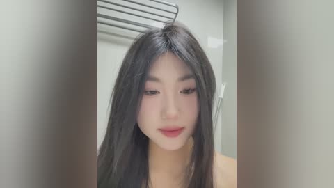 Media: Video of an East Asian woman with long, straight black hair and fair skin, wearing minimal makeup, standing in a bathroom with a silver towel rack in the background.