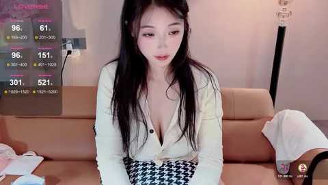 Media: A video of an East Asian woman with long black hair, fair skin, and small breasts, wearing a low-cut white cardigan and houndstooth skirt, sitting on a beige couch.