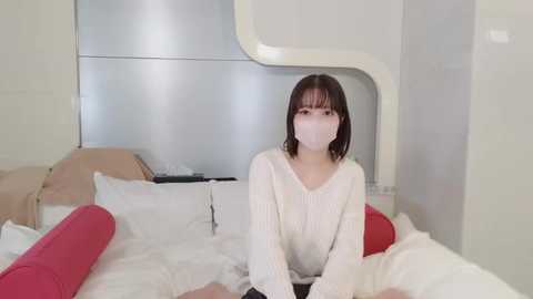 Media: A video of a young Asian woman with a light skin tone, wearing a white mask, white sweater, and seated on a bed with red and white sheets in a minimalist, modern room with a metallic headboard.