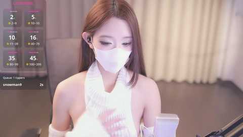 Media: Video of an Asian woman with light skin, long brown hair, wearing a white face mask, a white knit top, and a white wristband. She's sitting in an indoor setting with beige curtains in the background.