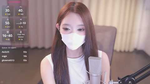 Media: Video of a young Asian woman with long brown hair, wearing a white mask and sleeveless top, sitting at a desk with a microphone, in a room with beige curtains.