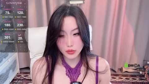 Media: Video of an Asian woman with long black hair, fair skin, and light makeup, wearing a purple lace top, sitting on a bed with a white pillow. Background shows a soft pink curtain.